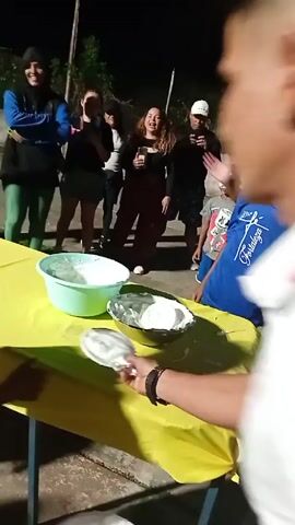 The video shows a group of people gathered outdoors at night, likely for a celebration or event. A yellow-covered table is in the foreground, with bowls and plates of white whipped cream or frosting on top. A hand reaches out and grabs a plate of the white topping. Several children are gathered around, and one boy is playfully smashed in the face with the topping. Other people are laughing and filming the event with their phones. The environment appears to be a paved outdoor area with trees and bushes in the background.