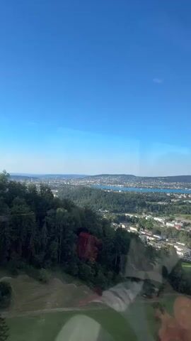 A vertical video begins with a clear blue sky. Below the sky is a wide expanse of green trees that cover a hillside, with a town visible in the distance at the bottom of the hill. In the background, a large body of water, likely a lake, stretches across the horizon, with more buildings dotting the landscape beyond the water. The video captures a panoramic view of a natural landscape integrated with a city.