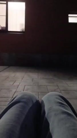 The video shows a first-person perspective of someone sitting down. The person's legs, clad in light blue sweatpants, are visible in the foreground. The background shifts between two scenes. In the first scene, the ground appears to be a paved surface, possibly concrete, with distinct lines suggesting tiles or slabs. In the second scene, the ground is dark and appears to be covered in dirt, gravel, and some debris. The lighting suggests an outdoor environment, possibly at dusk or night, with some artificial light sources visible in the distance in one of the background segments. There is no clear interaction or action taking place beyond the implied act of sitting.