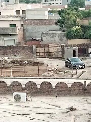The image captures an elevated view, likely from a rooftop, looking down onto a residential or semi-urban landscape in Dera Chahal, Pakistan.

In the immediate foreground, a white outdoor air conditioning unit is positioned on a flat, light-colored rooftop, accompanied by dark electrical wiring. Below this rooftop, a dusty, unpaved ground area is visible, partially enclosed by a simple, brown metal fence.

To the right of this ground area, a dark-colored SUV, possibly black, is parked. It faces slightly towards the left of the frame, with its front and side visible. Behind the vehicle and the fenced section, a substantial wooden gate, reinforced with metal cross-bracing, is set into a brick wall, suggesting an entrance to another property.

The background is dominated by a cluster of low-rise buildings, constructed from brick and concrete, characteristic of residential architecture. Many of these structures have flat roofs. Scattered among the buildings are a few green trees, adding touches of vegetation to the scene. The overall lighting suggests daytime, with diffused light indicating either an overcast sky, early morning, or late afternoon. There are no people visible, and no specific activity is taking place. No discernible text is visible on any objects or structures in the frame.