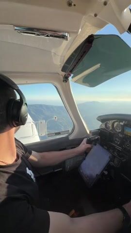 This is a first-person point-of-view video taken from the cockpit of a small airplane. The camera is mounted facing forward and slightly to the right, capturing the pilot's hands on the controls and the instrument panel. The pilot, a man wearing headphones and a dark t-shirt, is visible from the shoulder down, his left hand gripping the yoke and his right hand resting on the throttle. The instrument panel is filled with various gauges and screens, including a tablet mounted below the yoke displaying what appears to be navigation information. Sunlight streams in through the windshield, illuminating the cockpit and the sky outside, which is a clear blue with some clouds visible in the distance. The landscape outside the cockpit appears to be mountainous, with rolling hills and valleys stretching out to the horizon. The video shows the pilot's hands making minor adjustments to the controls, suggesting he is actively flying the aircraft. The overall impression is one of peaceful flight through a scenic landscape.