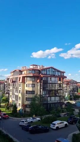 This vertical video captures a bright, sunny day in a modern residential complex in Romashkovo, Russia. The scene is dominated by several multi-story apartment buildings with distinctive red-tiled roofs and large, glass-fronted balconies. The architecture appears contemporary and well-maintained, suggesting a comfortable living environment.

The sky is a vivid blue, punctuated by fluffy white clouds, indicating pleasant weather. The time of day is clearly daytime, likely late morning or early afternoon, judging by the bright sunlight casting shadows. Lush green trees and bushes surround the buildings, adding a touch of nature to the urban landscape.

Several cars are visible parked on the road and in what appear to be designated parking areas. Among them, a red car is parked on the left, a white sedan and a white SUV are in the middle, and a black SUV is parked on the right. The cars suggest daily life is in motion within the complex. One of the buildings has a sign visible with "8K2" on it, which might be a building number or identifier.

There are a few people walking on the sidewalks, giving a sense of gentle activity and community. No particular event is being depicted; the video offers a serene glimpse into the everyday life of this suburban residential area. The overall mood conveyed is peaceful and serene, highlighted by the clear skies and the orderly appearance of the buildings and surroundings.