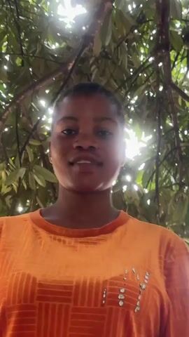 A young woman with braided hair and an orange shirt sticks out her tongue and smiles. She is outdoors, with trees and sunlight visible in the background.