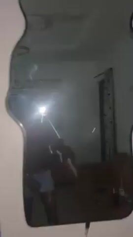 A person is standing in a dimly lit room, facing a mirror, and holding a phone with the flash on, obscuring their face. They are shirtless and wearing light-colored briefs. In the background, there is a window with bars, a door, a dresser, and a ceiling fan. The room appears to be a bedroom.