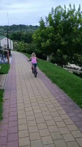 A child is riding a bicycle down a paved path in a park. The child is wearing a pink shirt and blue shorts. The path is made of brick pavers and is lined with grass on one side and a railing on the other. In the background, there are trees and a hill. The sky is cloudy.