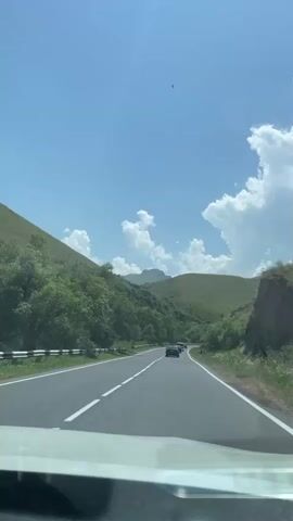 The video shows a POV shot from a car traveling on a two-lane road. The road is paved and has white lane markings. The car is moving through a mountainous or hilly area with green vegetation on both sides. There are several other cars on the road ahead. The sky is blue with scattered white clouds. The overall impression is of a scenic drive on a clear day.