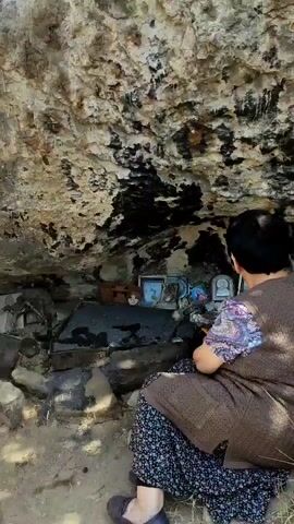 A woman kneels before a rock formation in what appears to be an outdoor setting. She is facing away from the camera and is wearing a brown vest over a patterned shirt and a dark skirt. In front of her, nestled into the rock, is a small shrine area. Several framed pictures or icons are arranged on a ledge. One picture appears to be of a religious figure. There are also some other small objects visible in the shrine. The rock face behind the shrine is rough and textured, with dark stains in places. The ground in front of the woman is covered with sand or dirt. The lighting suggests it is daytime.