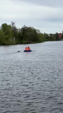 A long shot shows two people in a blue rowboat, each wearing bright orange life vests, rowing across a body of water. The water is textured with small waves. The rowboat is moving away from the camera. Along the banks are green trees and some houses are visible in the distance. The sky is overcast and gray.
