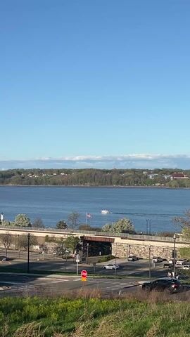 The video shows a sunny day over a body of water, likely a river or bay.  A small white boat with an American flag sails across the water from left to right. In the foreground is a bridge, with a few cars driving below.  A stop sign is visible near a grassy area.  In the background, there is a line of trees and buildings along the shore, with hills rising behind them.  The sky is blue with some scattered clouds.