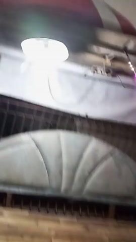 The video appears to be a short, shaky clip filmed at night or dusk, possibly in an outdoor setting like a market or festival. The first few seconds show a close-up of a bright light fixture, then pan upwards to a white awning with some lettering, and a glimpse of what looks like a padded seat or bench. The focus then shifts to strings of colorful lights, some resembling festive decorations, strung between trees and possibly some fabric awnings. Indonesian flags, red and white, are visible intermittently among the decorations. The background is dark, with indistinct shapes suggesting trees and perhaps other structures or people further away. The overall impression is of a lively, decorated outdoor space, possibly preparing for or in the midst of a celebration.