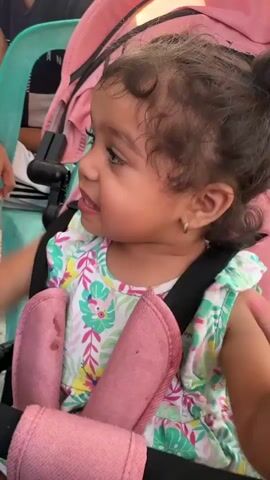 A close-up shot shows a young toddler sitting in a pink stroller. The toddler has dark curly hair pulled up into two buns and is wearing a white and floral print dress. The toddler is looking at the camera with a neutral expression. A person's hand, with a ring on their finger and yellow nail polish, reaches into the frame and gently touches the toddler's arm. The background is slightly blurred but appears to be an outdoor setting with greenery. The video is short, lasting only a few seconds, and shows a gentle interaction between the person and the toddler.