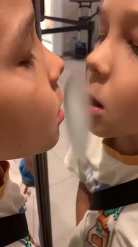 This media file features a young child looking at their reflection in a mirror. The child's face is close to the mirror, and they appear to be exhaling, creating a visible puff of mist or vapor between their lips and the reflective surface. The child is wearing a white t-shirt with a colorful graphic print and a black strap across their chest. The reflection shows the child's profile with their eyes closed or looking down.

The setting appears to be indoors, possibly a public space like a store or a waiting area, given the tiled floor, the presence of what looks like a stanchion or barrier in the background, and some equipment that could be related to retail or service. The lighting suggests it is daytime, but no specific time can be determined. The overall mood conveyed is one of curiosity and playfulness, as the child interacts with their own image. The location context of Surgut, Russia, is provided but not directly inferable from the visual elements in the media. There is no discernible text within the visible frame.