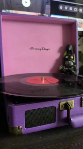The video shows a purple "Sunny Days" record player with a black vinyl record playing. The record has a red label in the center. A small, dark-colored statue of a meditating figure sits beside the record player. The record player is on a dark surface.