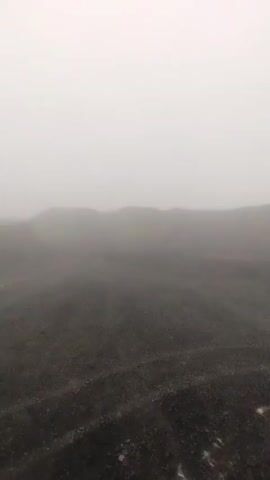 The video shows a desolate, foggy landscape. The ground is covered in dark, coarse gravel, with some larger rocks visible. In the distance, rolling hills are obscured by the thick fog, making it difficult to discern any specific features or vegetation. The sky is a uniform, pale gray, completely blending with the fog and the tops of the hills. There are no people, animals, or man-made objects visible in the frame. The overall impression is one of a stark and somewhat eerie environment, emphasizing the lack of visibility and the muted color palette.