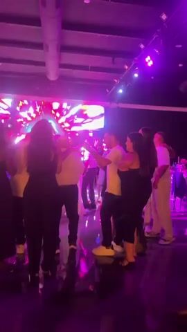 The video shows a group of people dancing in a dimly lit room. The room appears to be a club or event space, with a large screen displaying colorful abstract visuals at the back. There are also several spotlights illuminating the dance floor with changing colors, including pink, blue, and green. People are dressed casually, and some are holding drinks. The atmosphere is festive, with individuals moving to music that is not audible in the video. The camera captures various angles of the crowd, focusing on their interactions and movements on the dance floor.