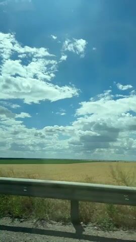 The video shows a view from a moving vehicle, looking out over a vast, rural landscape. The sky is bright blue with scattered white clouds. In the foreground, there is a metal guardrail. Beyond the guardrail, there are fields of golden wheat and green crops, stretching to the horizon. The sun is shining, casting a bright light on the scene. The wind is blowing, causing some of the green foliage to sway.