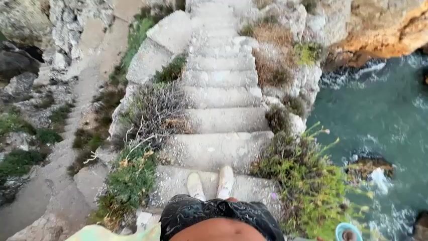 The video begins with a first-person perspective looking down a set of rough, concrete stairs carved into a rocky cliffside. The stairs are worn and uneven, with sparse vegetation growing around them. The ocean, with its turquoise-green water, is visible to the right of the stairs, crashing against the rocks below. As the video progresses, the camera moves downwards, following the stairs. The perspective becomes more dynamic, showing a rapid descent. The scene then pans out, revealing the grandeur of the coastal landscape. Towering cliffs flank a narrow inlet of the sea, with the water appearing calm and inviting in the distance. The overall impression is one of adventure and exploration, with the viewer being guided down a rugged path towards the water.