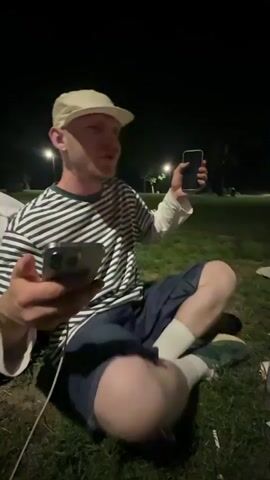 The video shows a man sitting cross-legged on a grassy area at night. He is wearing a white baseball cap, a striped long-sleeved shirt, blue shorts, and white socks. He holds two smartphones in his hands and looks at one of them. Another person sits nearby, holding a can. In the background are dim streetlights. There is a bag and some other objects on the grass around the people.