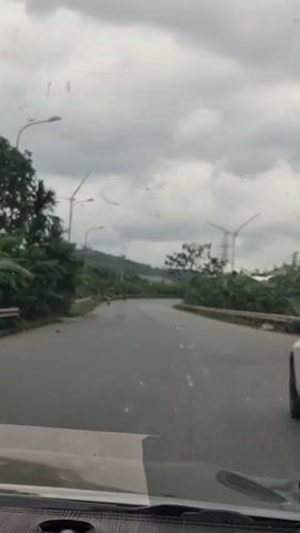 The video shows a view from inside a vehicle, likely a car, driving on a paved road. The sky is overcast with heavy clouds. On either side of the road are lush green trees and vegetation, some appearing tropical. In the distance, there are hills and what appear to be wind turbines, suggesting a rural or semi-rural landscape. There are also power lines visible. Two individuals on motorcycles are seen riding ahead on the road. The road itself has some potholes and painted lane markings. The dashboard of the vehicle is visible in the foreground.