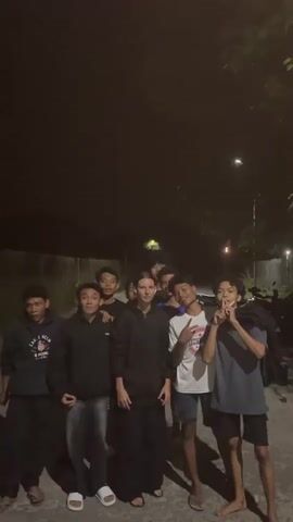 A group of young people, appearing to be mostly male with one female in the center, pose for a video in a dimly lit outdoor environment at night. The background consists of a fence, some trees, and a streetlamp. The individuals are making various hand gestures and facial expressions, seemingly in a playful or celebratory manner. Some are smiling and looking directly at the camera, while others are engaging with each other. One individual is wearing a black hooded sweatshirt, another a white t-shirt with graphics, and another a blue t-shirt. The female in the center is wearing a black hooded garment and dark pants. The ground is paved, and there are shadows cast by the individuals and trees. The overall atmosphere appears to be informal and energetic.