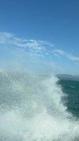 A video captures a close-up view of ocean waves crashing and splashing against the camera. The water appears turbulent and frothy, with droplets suspended in the air. In the background, the blue sky is visible with scattered white clouds. Further in the distance, a hazy coastline with buildings can be seen. The perspective suggests the camera is either on a moving boat or very close to the water's surface. The scene conveys a sense of movement and the power of the ocean.