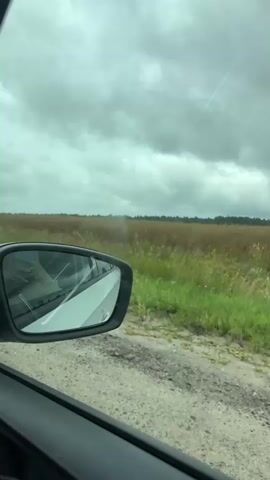 The video shows a view from inside a car, looking out the side window. The car is driving on a gravel road next to a field of tall grass and some trees in the distance. The sky is cloudy. A side mirror of the car is visible in the foreground.