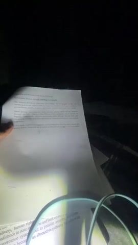 In a dimly lit environment, a hand is holding a white sheet of paper illuminated by a bright light source, seemingly a flashlight. The paper is covered in printed text, possibly a document or article. The hand is moving the paper slightly, adjusting its position in relation to the light.  A circular object with a wire frame, possibly a magnifier, is also present in the frame, partially obscuring the text at the bottom of the page. The background is mostly black, suggesting a dark room or outdoor setting at night. The focus is primarily on the paper and the act of reading or examining it under low light conditions.