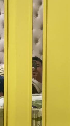A person is lying in bed and taking a selfie in a closet mirror. The closet doors are yellow and are slightly ajar, revealing a sliver of the mirror. The person's face is visible in the mirror, holding a phone to take the photo. The bed has a tufted headboard and patterned bedding.