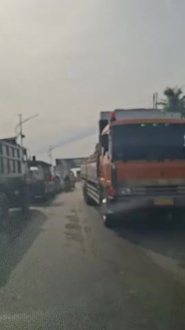 The video is shot from a moving vehicle on a road. It shows a traffic jam with several trucks and some motorcycles. The road appears wet, and the sky is overcast. The trucks are of various sizes and colors, including a large orange truck in the foreground loaded with what appear to be sacks or bags. In the background, there is an overhead structure, possibly a bridge or an overpass, and more traffic. Some people are visible on the motorcycles, and some figures are standing near the trucks. The overall scene depicts a congested road situation.