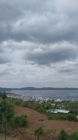 This is a landscape video taken from a hillside, looking down towards a coastal town and the ocean. The sky is filled with dramatic, heavy clouds, suggesting an overcast or possibly stormy day. In the foreground, there's a grassy slope with dry patches and some scattered green bushes and trees. Below the slope, a cluster of buildings forming a town is visible, with white and red roofs. Beyond the town, the ocean stretches out, dotted with numerous small boats or fishing vessels, creating a textured surface on the water. In the distance, a line of hills or mountains can be seen on the horizon. The overall impression is a somewhat moody and natural scene with human settlement encroaching on the coastline.