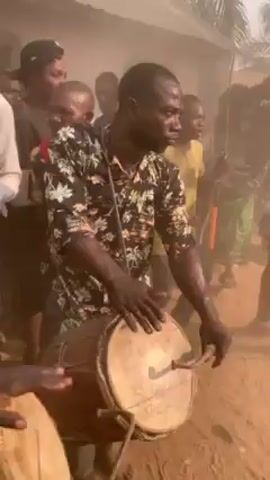 The video shows a group of people gathered in a dusty outdoor setting, with several individuals playing drums. The primary focus is on a man in a black and white floral shirt playing a light-colored drum with a stick. Other people are visible in the background, some appear to be singing or chanting. The atmosphere seems lively and potentially ceremonial. Dust or smoke is visible in the air.