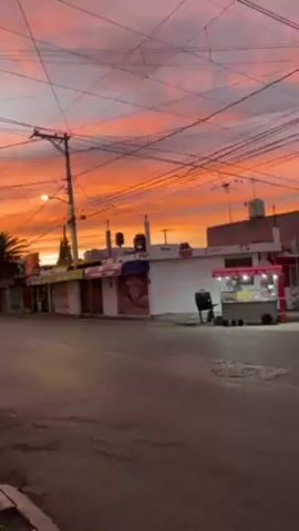 The video shows a street scene at sunset.  The sky is ablaze with orange and pink hues. Buildings line the street, including a storefront with a colorful awning and a food vendor cart. Utility poles crisscross the sky with a tangle of wires.  A person partially obscured by a dark frame is visible at one point.  Some cars drive by in the background.