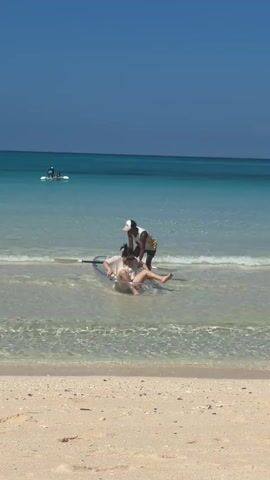 A clear, sunny day at the beach is captured. The water is a vibrant turquoise, gradually deepening to a rich blue on the horizon. The sand in the foreground is light and granular. In the middle ground, two individuals are interacting with a clear kayak near the water's edge. One person is seated in the kayak, appearing to be adjusting or getting settled. The other person, standing behind the kayak, is leaning over and appears to be assisting them. Their actions suggest they are either preparing to launch or have just returned. Further out in the water, another person is visible on a paddleboard, standing upright and paddling. The overall scene conveys a relaxed and active day by the sea.