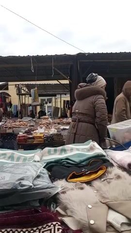 The video shows an outdoor market with various stalls under a covered area. People are walking around, some browsing goods on display. The stalls appear to be selling a mix of items, including food items like oil, nuts, and dried goods, as well as textiles such as fabrics and scarves. The sky is overcast. Some people are carrying shopping bags. The ground appears to be a mix of concrete and dirt. The overall atmosphere suggests a busy local market.
