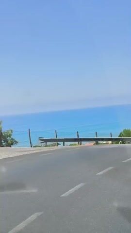 This is a video taken from inside a moving vehicle. The view is through the passenger side window, and the camera is looking out at a coastal landscape. The ocean is visible in the background, a bright blue under a clear sky. In the mid-ground, there is a roadside barrier, followed by a stretch of green vegetation and several buildings with red-tiled roofs. A few trees are also present. The road itself, a gray asphalt surface, occupies the foreground, with white lane markings visible. The driver's side rearview mirror is also visible in the frame, reflecting the sky and some trees.