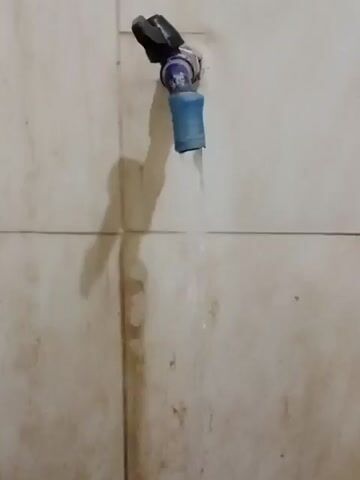A faucet is seen dripping water into a blue bucket below, on a tiled wall. The faucet has a black handle and a blue plastic extension. The tiles are off-white and appear dirty, with visible water stains running down the wall. The bucket is partially visible at the bottom of the frame and is filled with water. The lighting is dim, casting shadows on the wall.
