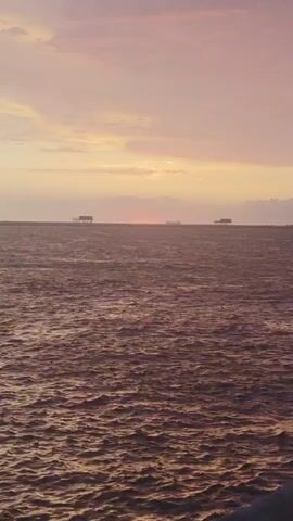 The video shows a wide shot of a choppy, reddish-brown sea at sunset. Three distant, dark rectangular structures, possibly oil rigs or barges, are visible on the horizon. The sky above is a gradient of pale pink and yellow, with scattered clouds. A small, dark speck, likely a bird, is present in the upper right quadrant of the sky. The sun is a faint reddish glow on the horizon, partially obscured by clouds. The lighting suggests it is either dawn or dusk.