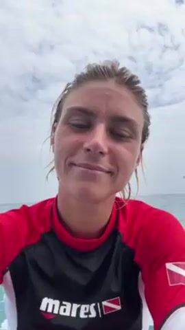 The video shows a woman in a red and black wetsuit sitting on a boat, facing the camera with a slight smile. Her wet hair is pulled back, and she rests her chin on her hand. The ocean and a cloudy sky are visible in the background. The wetsuit has "mares" printed on the front and a diving flag on the sleeve. The boat appears to be moving.