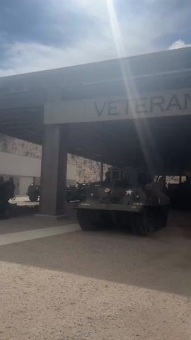 The video shows a collection of military tanks and armored vehicles parked under a covered pavilion at a Veterans' memorial. The vehicles are painted green and bear the iconic white star insignia, a symbol of U.S. military vehicles. The pavilion provides shade, and the background reveals a mountainous landscape under a partly cloudy sky. The ground is gravel, and the atmosphere suggests a memorial or museum dedicated to military history.