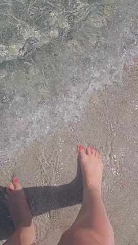 The video shows a first-person perspective of someone walking in shallow, clear water on a sandy beach. The water is transparent, revealing the sandy bottom and small ripples on the surface. The person's feet are visible as they step into the water and move forward. The light reflects off the water, creating shimmering patterns. The environment appears to be a calm, sunny day at the beach.