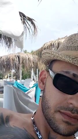 The video begins with a close-up selfie of a man wearing a straw hat and sunglasses. He has a beard and a silver chain around his neck, and tattoos are visible on his shoulder. The background shows a beach setting with palm umbrellas and white canopies. The scene then shifts to a wider shot revealing a beach with a pebble shore, calm water, and distant hills. Two iced drinks with straws are placed on a patterned mat in the foreground, suggesting a relaxed atmosphere. The camera pans slightly to show more of the beach huts and umbrellas. Finally, the view focuses on a wooden post with the number 504 painted in yellow, further emphasizing the beach resort or club setting. The overall environment is a sunny day at a beach resort with amenities for relaxation.