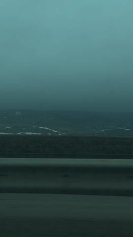 The video shows a view from a moving car on a highway. The sky is a muted, overcast turquoise. In the distance, hills are visible with patches of snow. A grey guardrail runs along the side of the road. Music can be heard in the background.