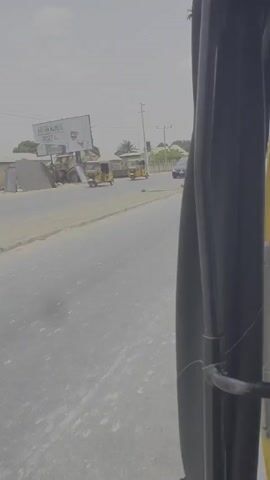 The video shows a view from inside a yellow auto-rickshaw, commonly known as a "keke," traveling on a paved road.  The windshield wiper is not moving. Another yellow keke is ahead. The seat in front of the camera is dark purple and patched with tape.  A person wearing a gray shirt and lime green vest is driving.  The surrounding environment includes electric poles, buildings, trees, and a billboard, visible under a bright, overcast sky.