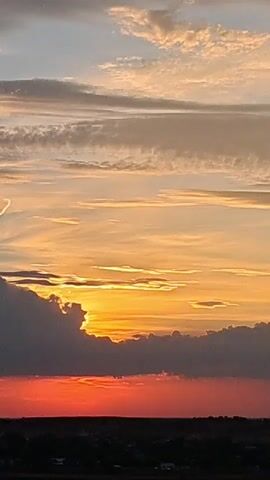 This is a vertical video of a sunset. The sky is filled with clouds of various colors, including orange, yellow, pink, and gray. The sun is setting behind a dark cloud formation, casting a bright orange glow. The lower portion of the video shows a dark landscape, likely a horizon line with trees and possibly some buildings. There are no people or explicit objects visible besides the natural elements. The video is static, with no movement other than subtle shifts in the cloud formations.