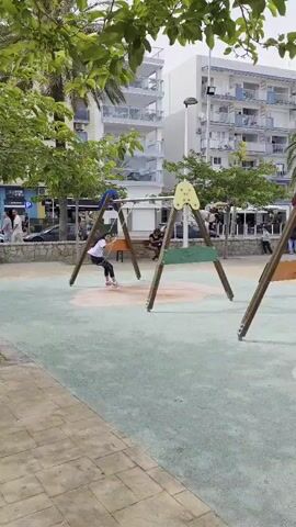 The scene depicts a playground with three swings. Two individuals, a female and a male, are seen swinging on two of the swings. A young boy in a black jacket and shorts walks around the playground, holding a red soccer ball. The playground has a green padded surface, and is surrounded by trees and buildings. There are other people walking in the background. The weather appears to be sunny.