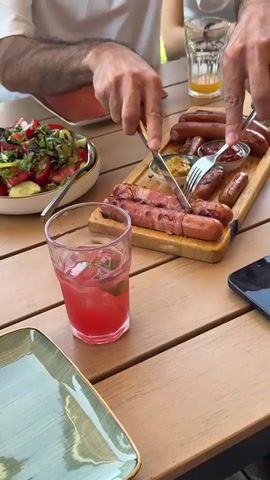 The video shows a close-up of a wooden table laden with food. In the foreground, a glass of red liquid with ice and mint sits beside a green plate with a wrapped sausage. To the right, a person’s hands are visible using a knife and fork to cut a sausage from a wooden platter that holds several sausages, some wrapped in bacon, along with small dishes of mustard and ketchup. Behind the platter, a bowl of salad with tomatoes, cucumbers, and greens is visible. Another person’s arms and shoulders are in the upper left, also appearing to be eating. The table appears to be outdoors, as suggested by the natural light and the presence of a glass of orange liquid and possibly another drink in the background. The overall scene depicts a meal being shared.