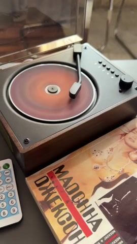 A close-up shot shows a record player with a spinning record. A remote control and a book are placed next to it. The book is titled "Madonna & Jackson." The record player has a wooden base and a metal top. The remote control is white with blue buttons. The book is open and shows a picture of Madonna on the cover. The background is blurry, suggesting an indoor setting.