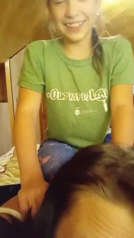 The video shows two people, likely a mother and daughter, in an indoor setting with a wooden-paneled ceiling. The foreground is dominated by the head of the person filming, with dark hair and an upward-facing gaze. Behind them, a young girl with long brown hair tied in a ponytail is seated. She is wearing a green t-shirt with white text and blue jeans with a butterfly pattern. The girl is smiling and looking down at the person filming her, with her hands near their head. The lighting suggests it is daytime, and the setting appears to be a cozy room, possibly a bedroom or living area in a home. The overall mood seems lighthearted and playful.