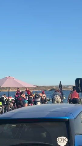 A group of people are on stationary bicycles outdoors, with a body of water and land in the background. The people are dressed in athletic wear, and some are wearing helmets. There are umbrellas providing shade. The scene appears to be an event or competition.
