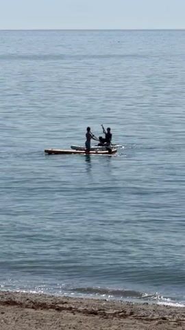 The video shows two people on a paddleboard in the ocean. The paddleboard is orange and white. The water is blue and there are waves crashing on the shore in the foreground. The sky is clear and blue. The two people are standing on the paddleboard and appear to be paddling. They are both wearing dark clothing. The person in the front is facing away from the camera, while the person in the back is facing towards the camera. The paddleboard is moving from left to right in the frame. The video is 4 seconds long.